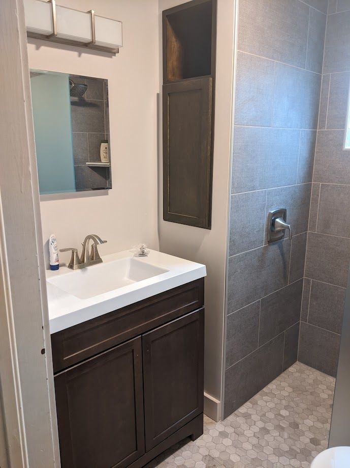 A bathroom featuring a dark wood vanity with a white sink, a mirror, wall lights, a tall storage unit, and a tiled shower.