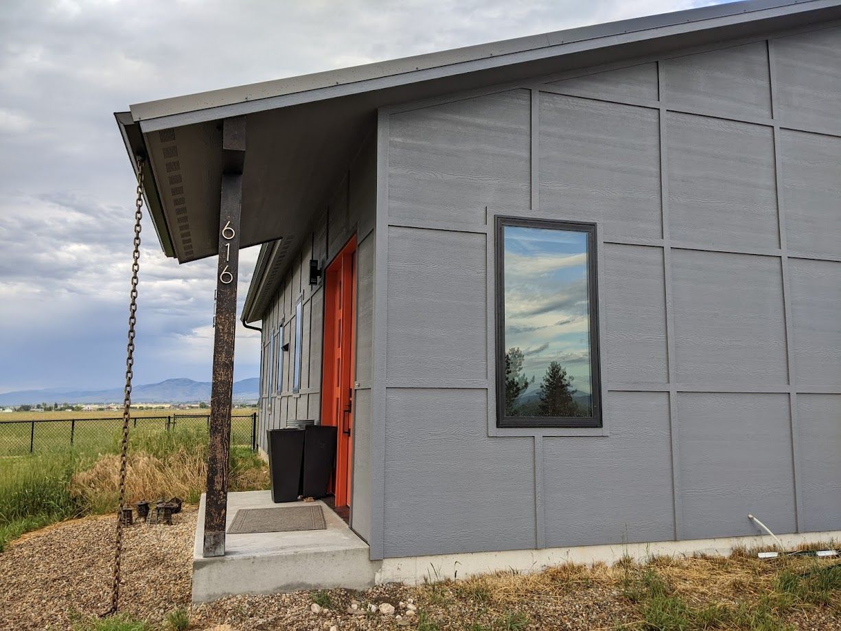 Gray modern home with vertical siding, a bright orange door, a large window, and a rustic wooden support post.