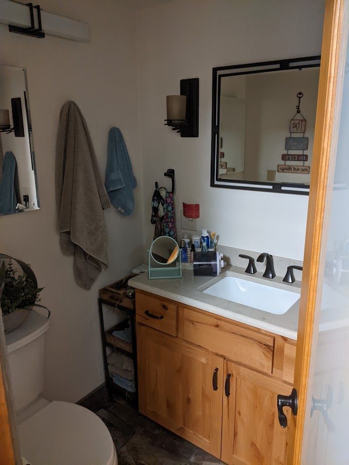 A bathroom vanity with a light wood cabinet, white sink, wall-mounted mirror, and wall sconce next to a toilet.