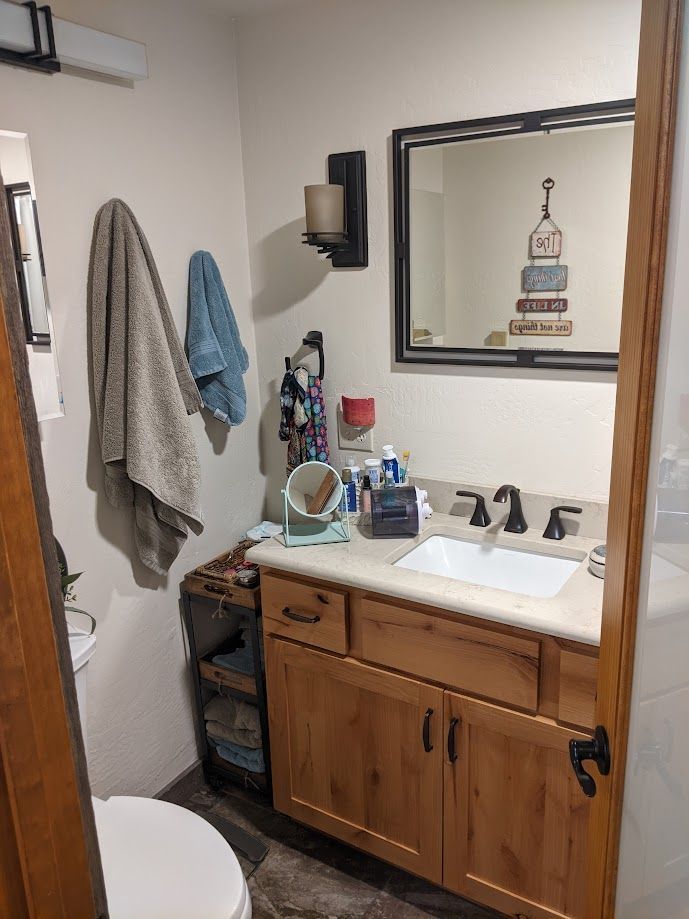 A small bathroom with a wooden vanity, a white sink, a mirror with decorative signs, and towels hanging on the wall.