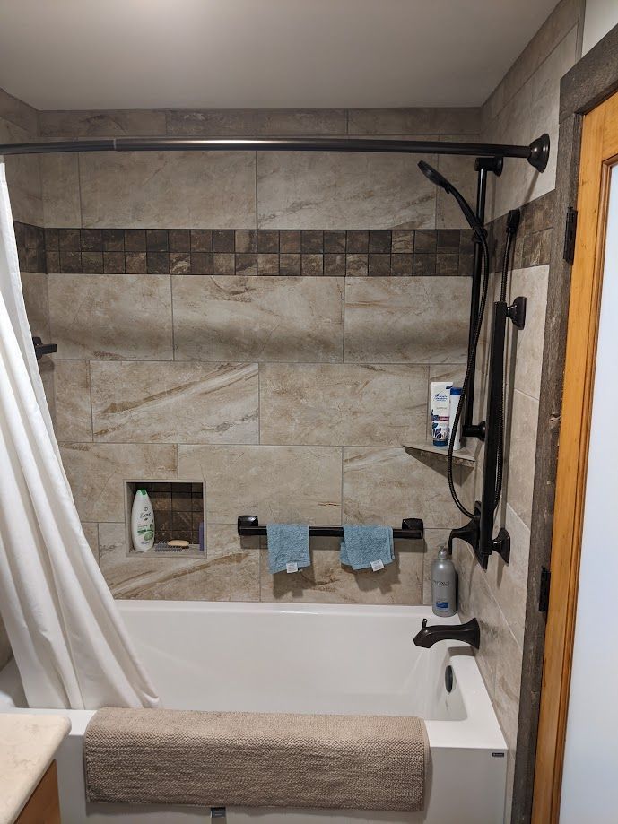 A bathroom shower-tub combo with tan tiled walls, a small recessed shelf, a black shower head, and a white curtain.