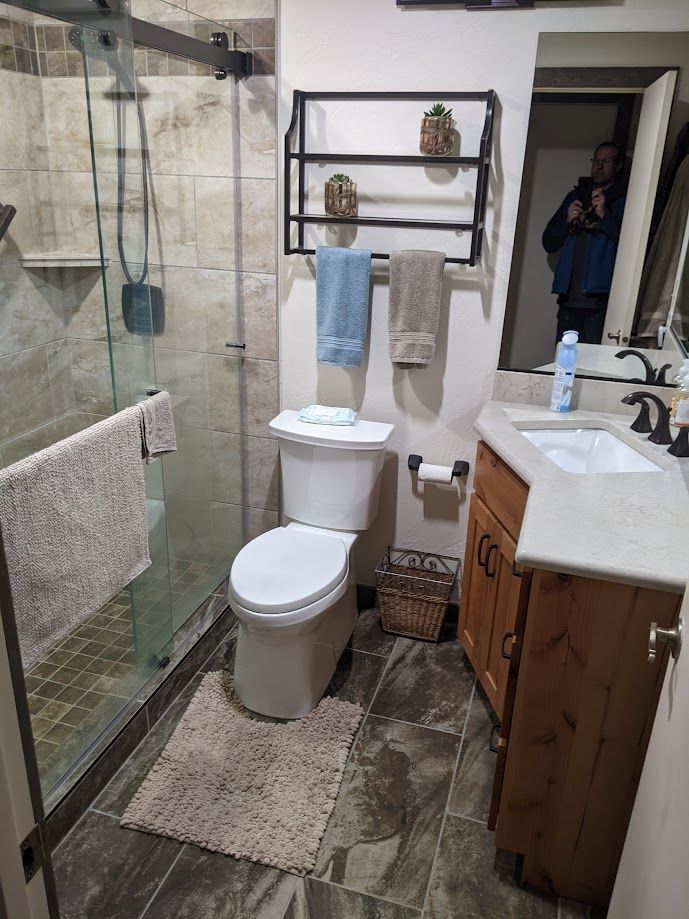 A bathroom with a walk-in glass shower, toilet, wall shelving, and a wood vanity with a white sink.