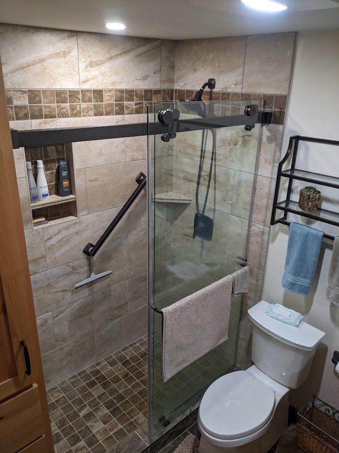 A small walk-in shower with stone tile, a sliding glass door, a black grab bar, and a niche, beside a toilet.