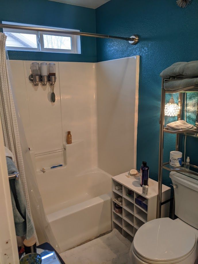 A bathroom with blue walls, a white shower-tub surround, a small white cubby organizer, and a toilet with a shelf above it.
