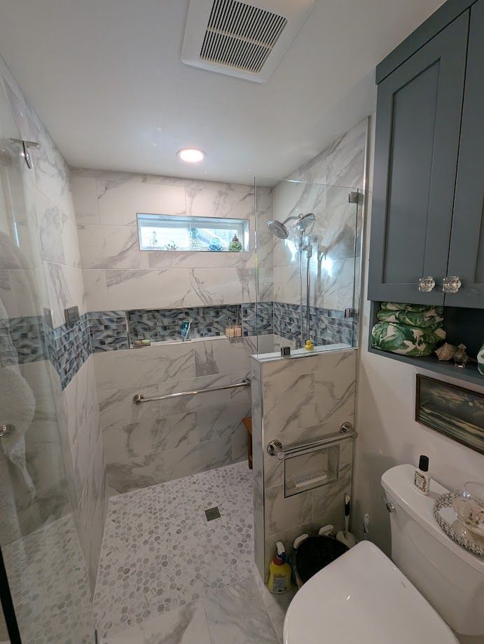 A bathroom with a glass-enclosed walk-in marble shower featuring blue tile accents and a toilet next to gray cabinetry.