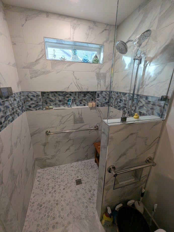An accessible walk-in shower with marble-style tiles, a bench, grab bars, a handheld showerhead, and a small window.