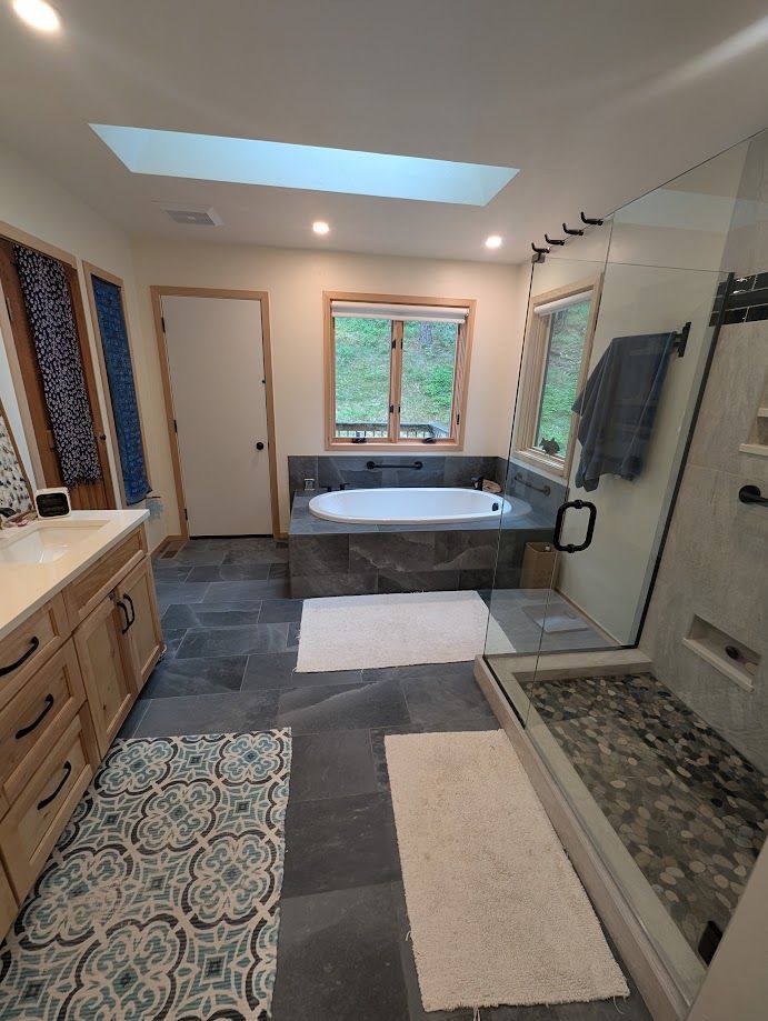 A modern bathroom with slate tile floors, a jetted soaking tub, a large glass-enclosed shower, and a wooden vanity.