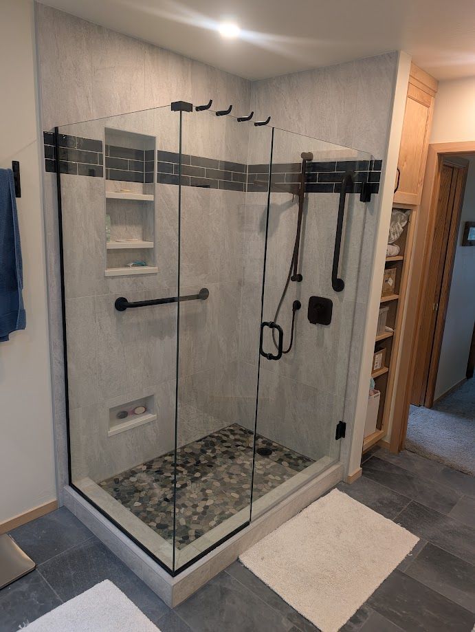 A modern shower with glass walls, black fixtures, a pebble floor, a built-in shelf, and grab bars in a light gray bathroom.