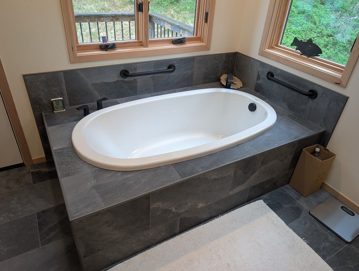White oval bathtub set in a dark gray tiled deck with two black safety grab bars and windows in a bathroom.