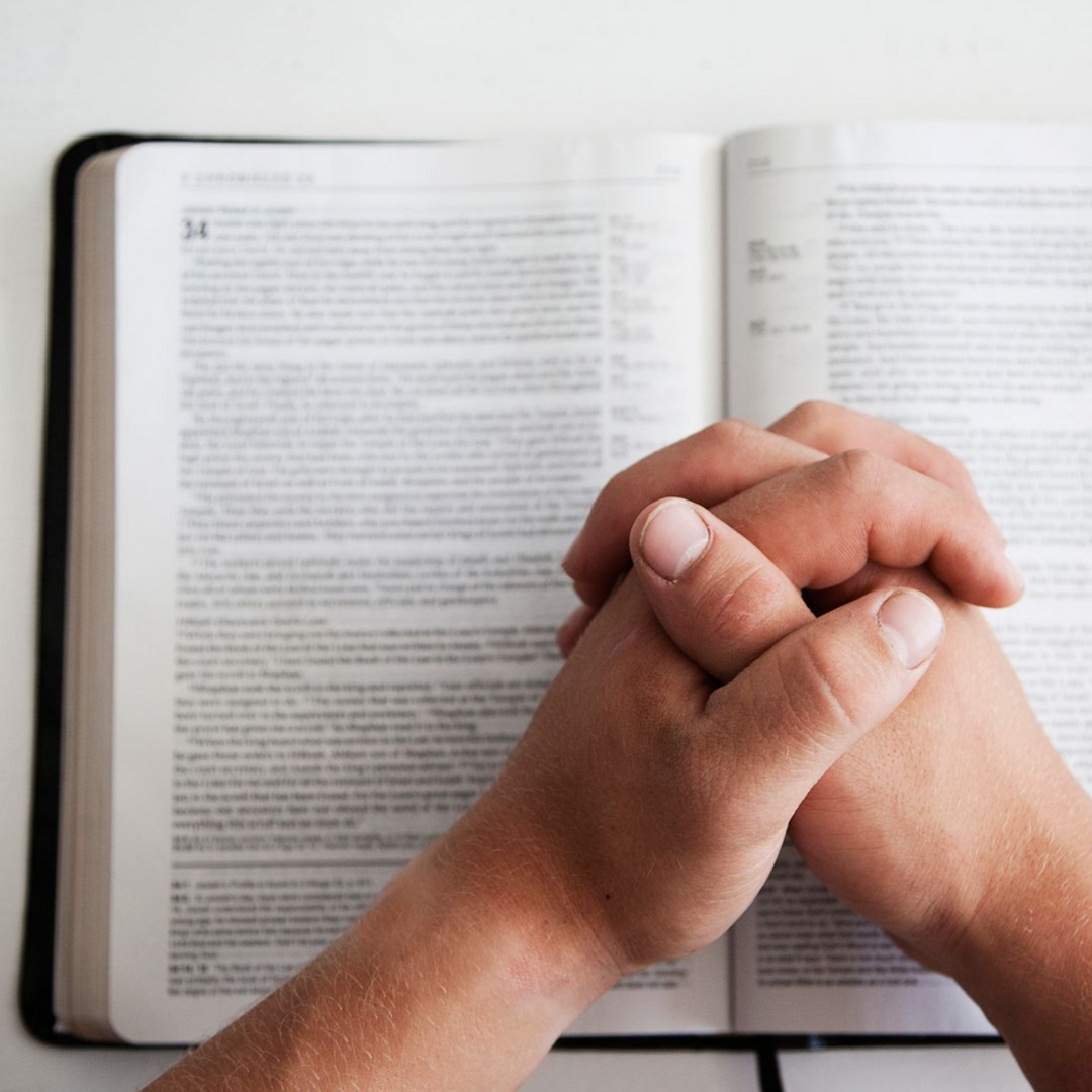 Hands folded in prayer resting on an open book, likely a Bible.