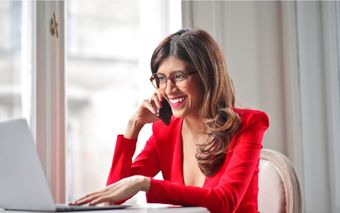 Una mujer con una chaqueta roja está hablando por un teléfono celular mientras usa una computadora portátil.