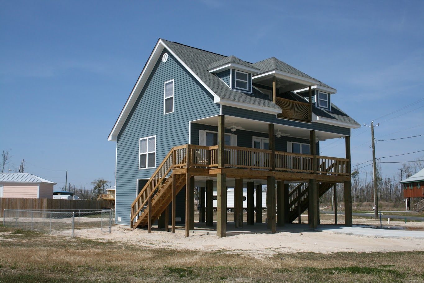 A blue house with a wooden deck is sitting on top of a sandy field.