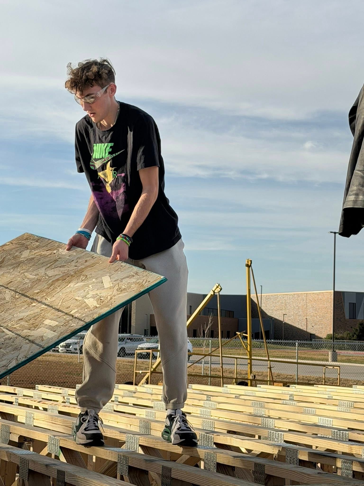 A person in safety glasses holds a wooden panel while standing on an unfinished floor frame outdoors.