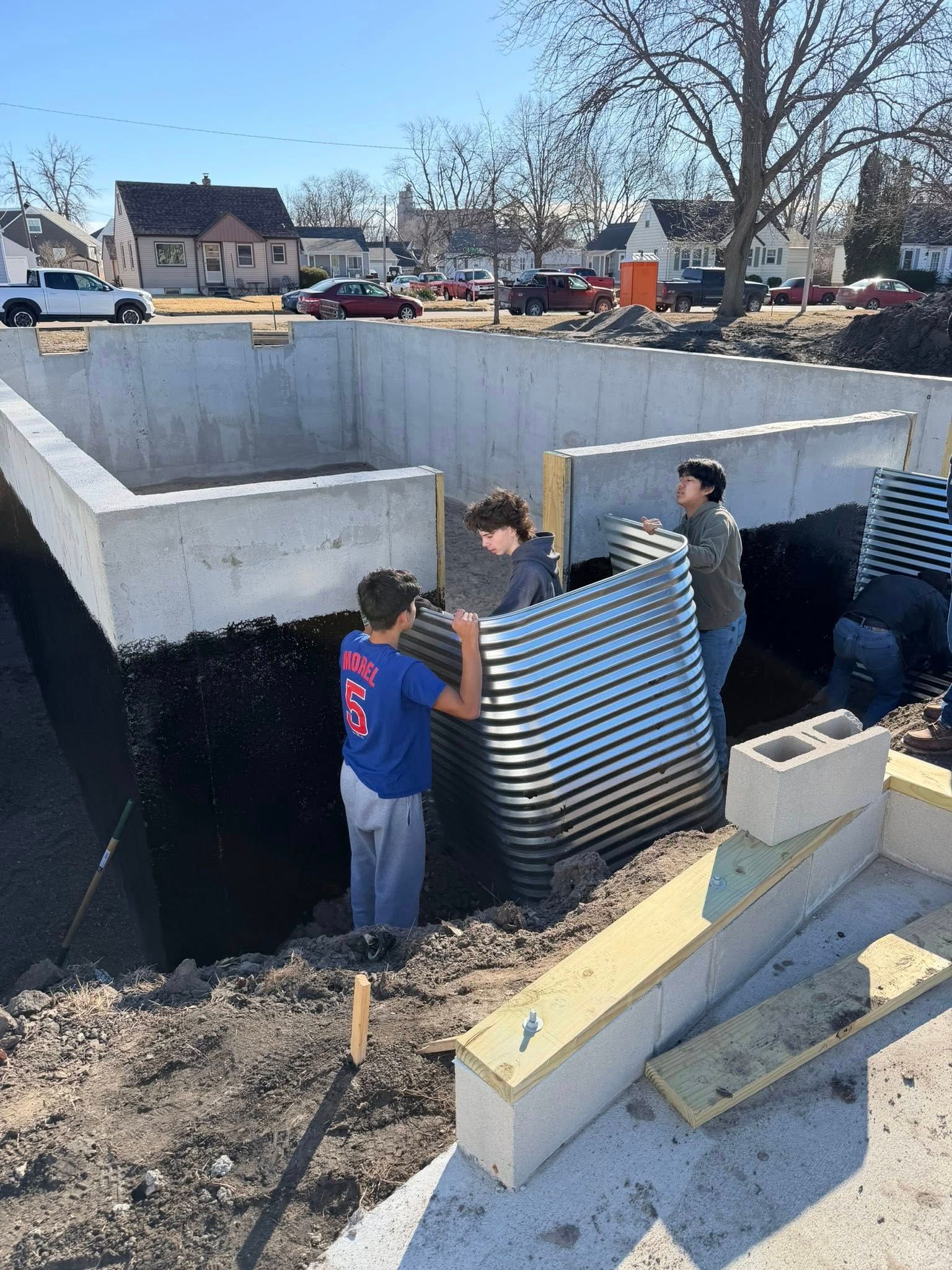 Three people install a corrugated metal window well against a concrete foundation wall on a construction site.