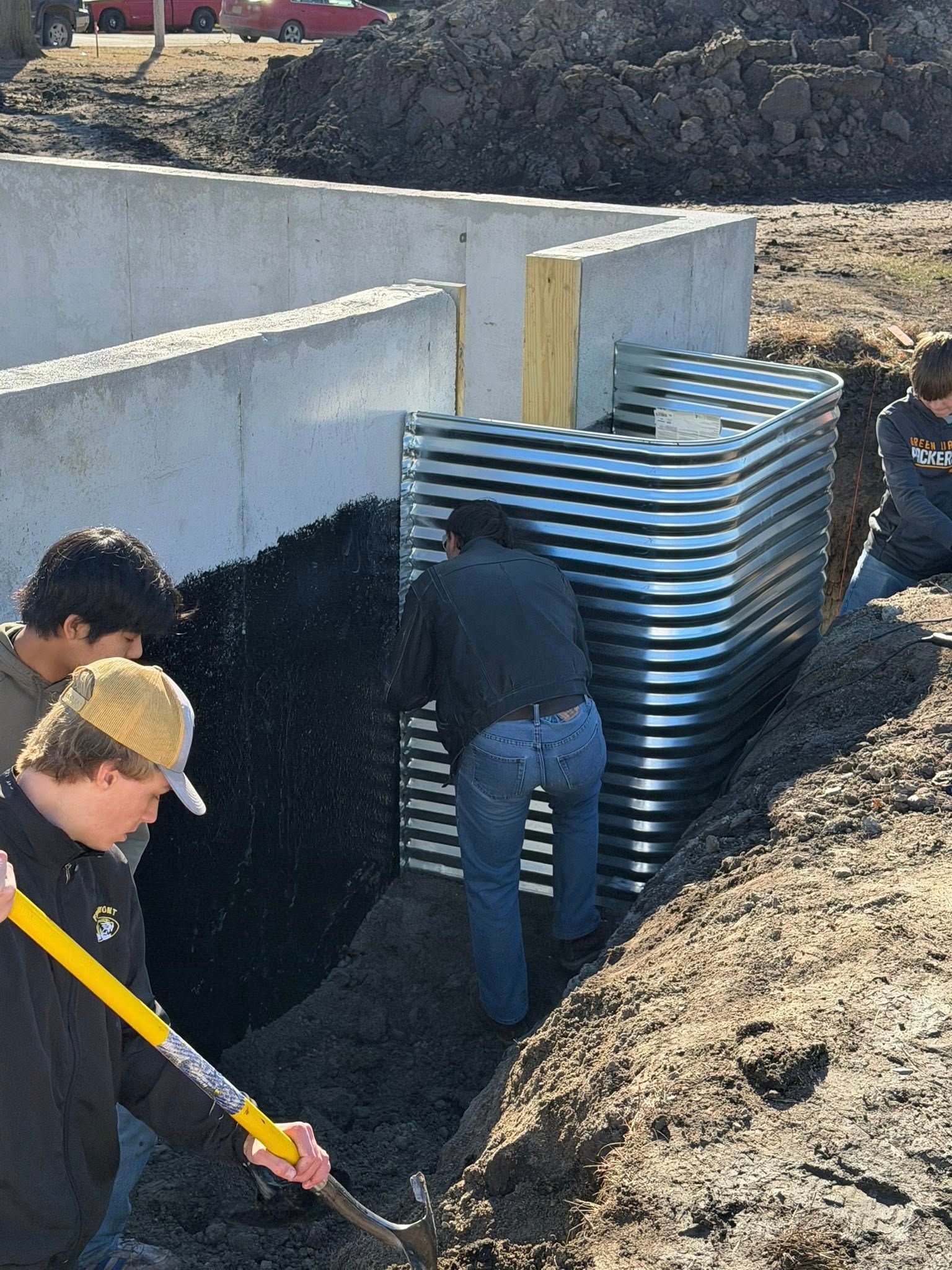 Three people installing a corrugated metal window well against a concrete foundation wall on a construction site.