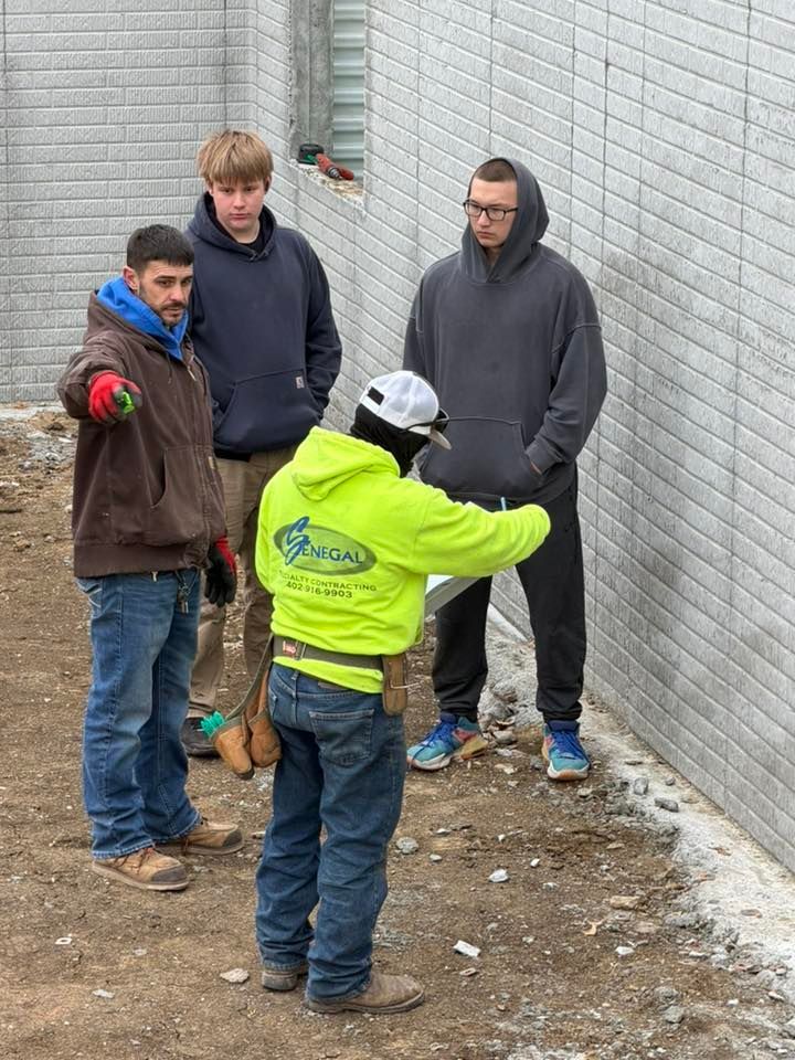 Four people standing on dirt outside a gray brick wall, with one person in a bright yellow shirt pointing toward the wall.