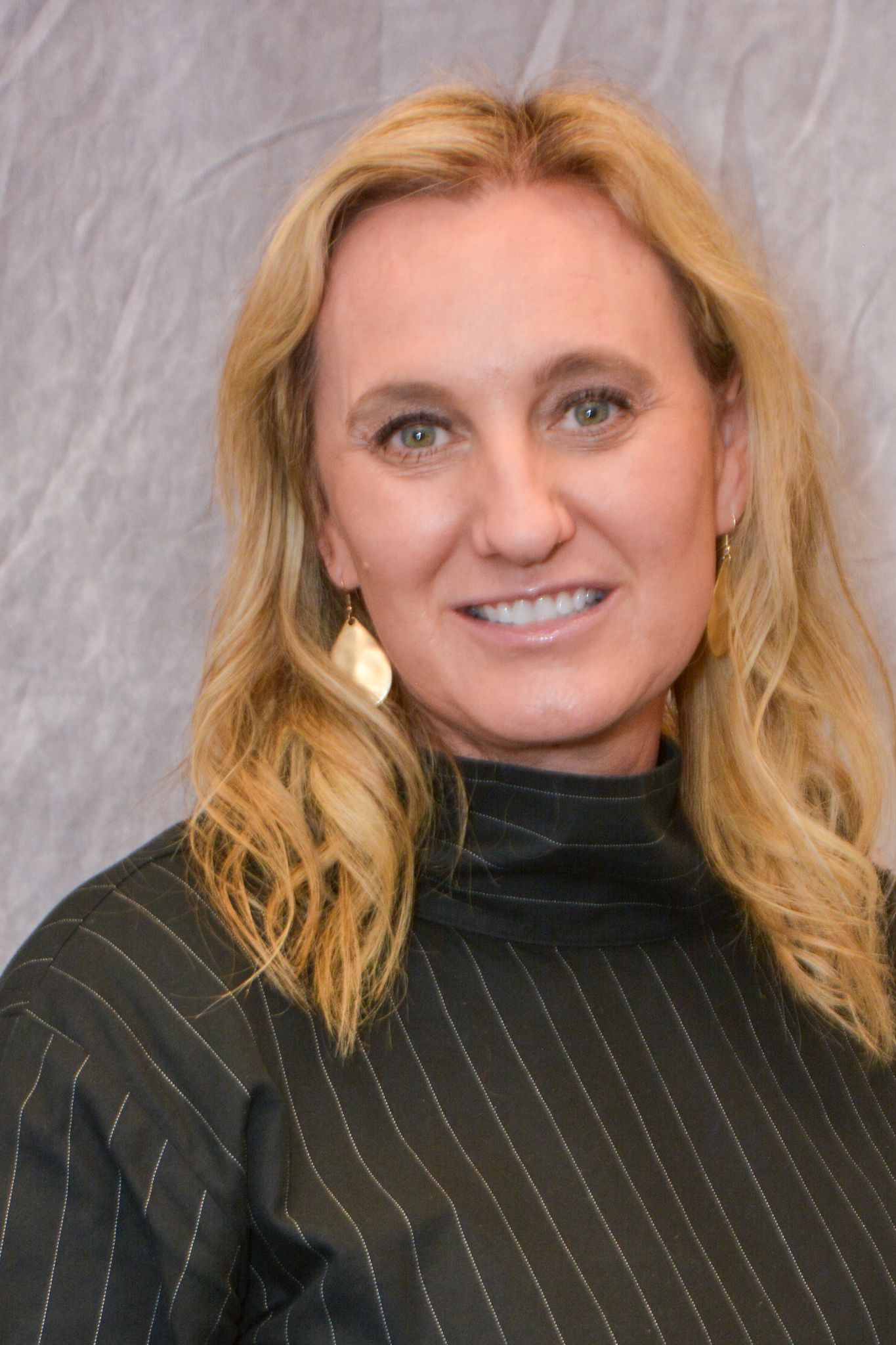 Headshot of a smiling person with wavy blonde hair wearing earrings and a dark top against a light gray background.