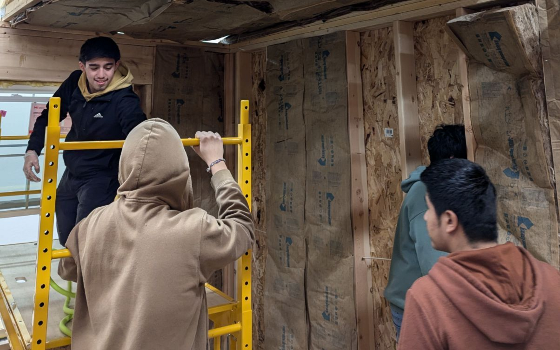 Four people work on constructing an interior wall, installing insulation between wooden studs.