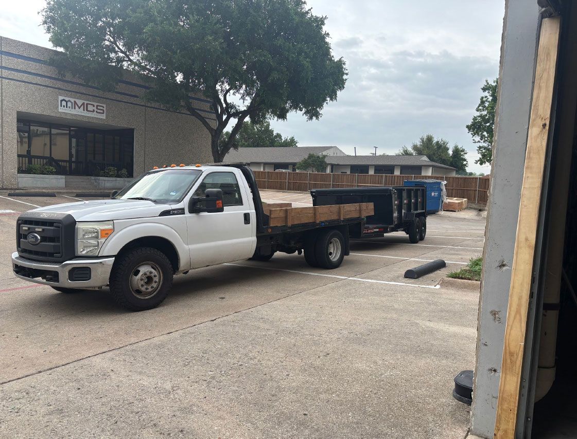 White flatbed truck parked by a building on a gravel lot, with a blue dumpster behind it.