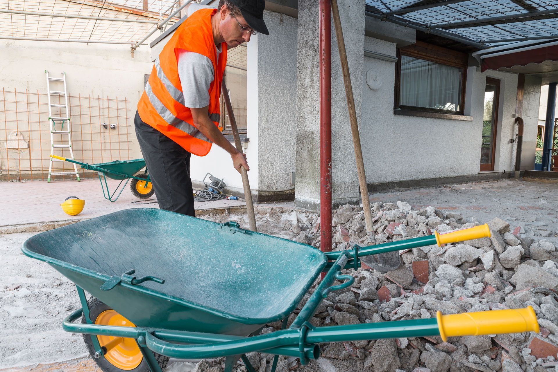 Worker in orange safety vest shovels rubble into a green wheelbarrow at a construction site.