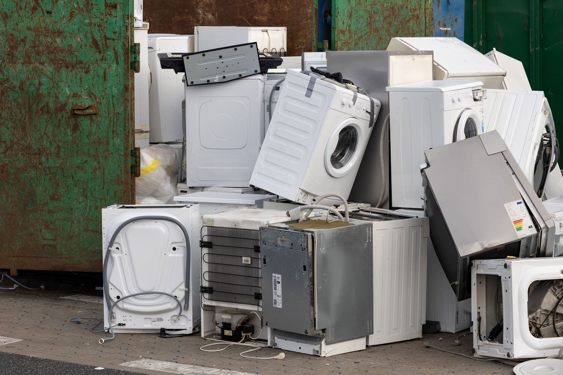 Pile of discarded white appliances and metal scrap beside a rusted green dumpster.