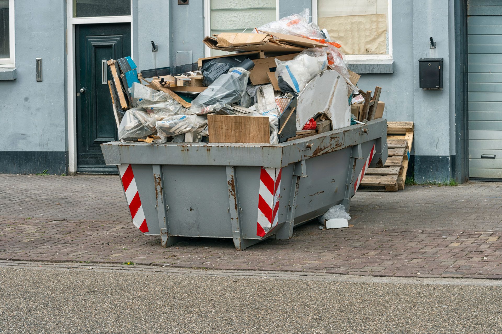 Large gray dumpster overflowing with cardboard and debris on a street outside a building