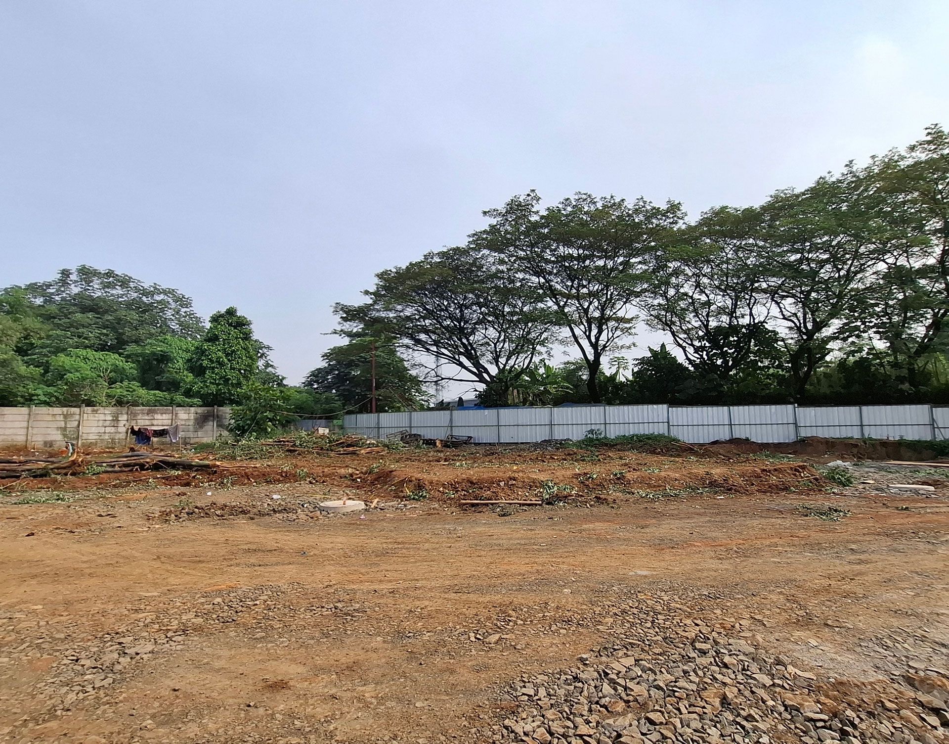 Cleared dirt lot with rubble, white fence, and trees under a cloudy sky