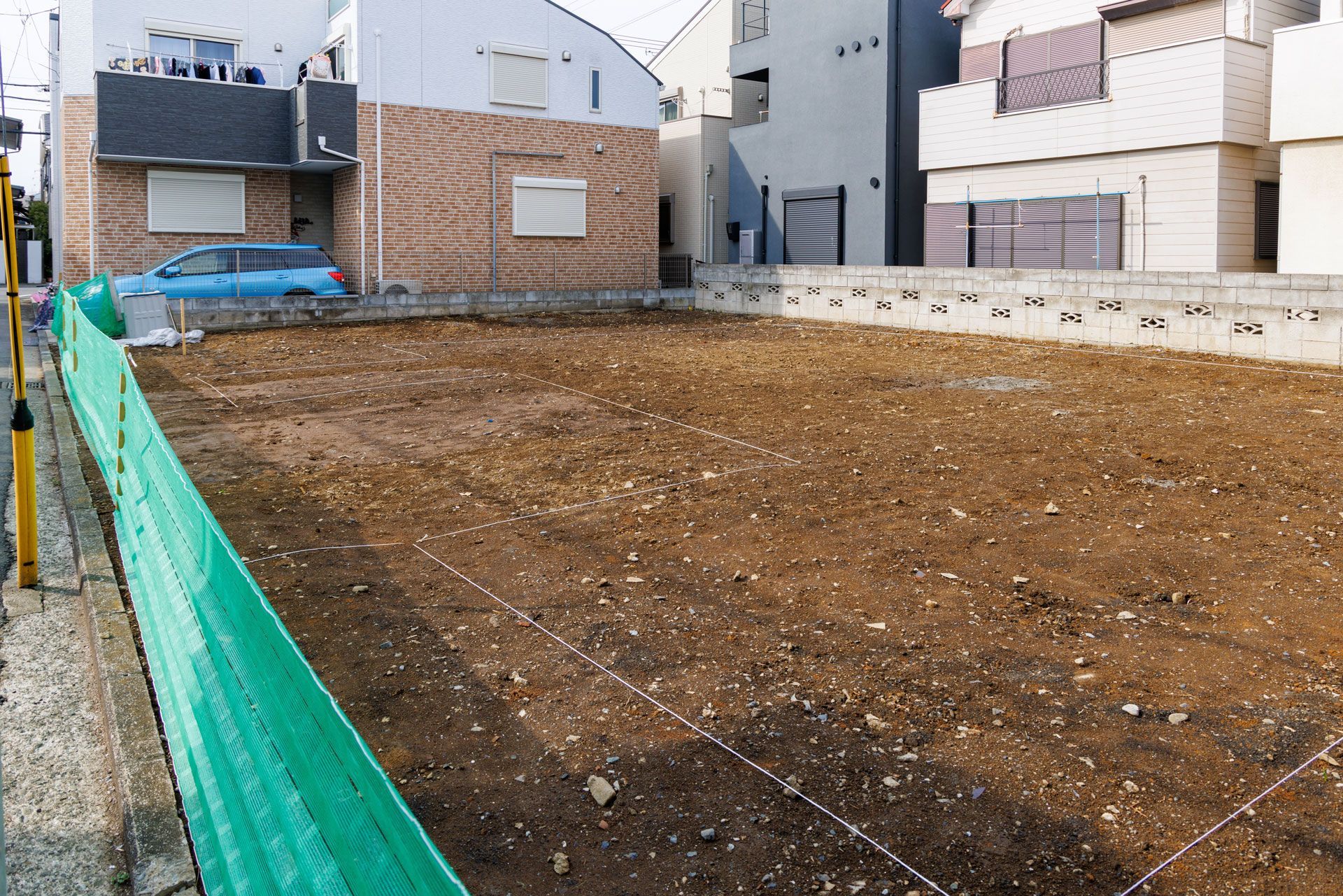 Empty dirt lot beside residential buildings, bordered by green mesh fencing and concrete walls