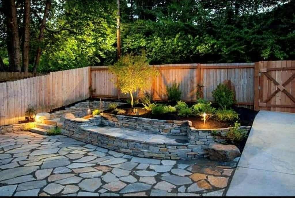 Stone patio with retaining wall, small trees, and landscape lighting next to a wooden fence.