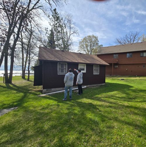 Two people stand beside a small dark cabin on a grassy lakeside lawn.