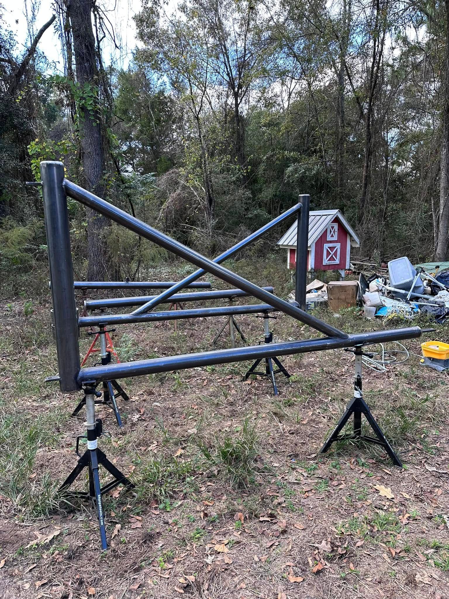 Metal frame structure on tripods outdoors, with small red building in the background.