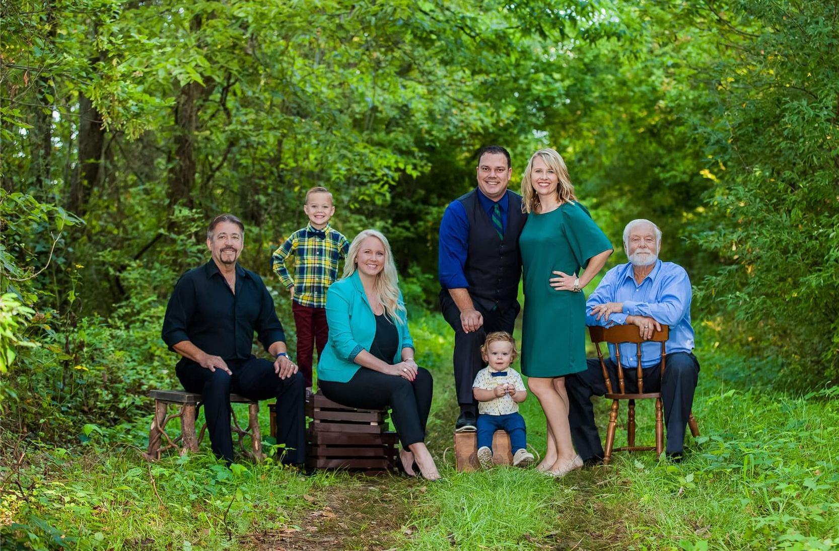 Family posing outdoors; adults seated on chairs, two children standing, and one held, all smiling amidst greenery.