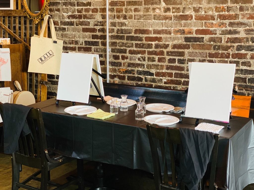 Table set up for painting with canvases, supplies, and chairs in front of a brick wall.