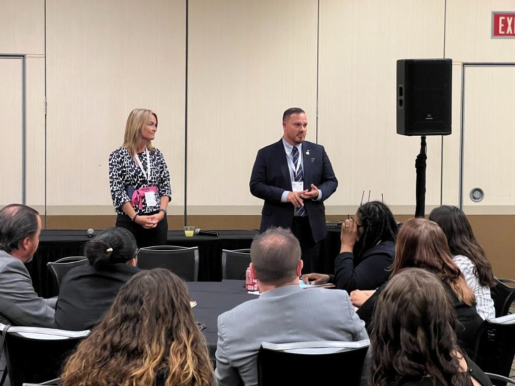 Two speakers addressing an audience seated at a table. A speaker stands next to a tall speaker.