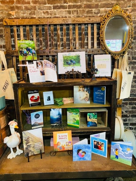 Books and cards displayed on a wooden shelf in front of a brick wall, with a gold mirror and goose figurine.