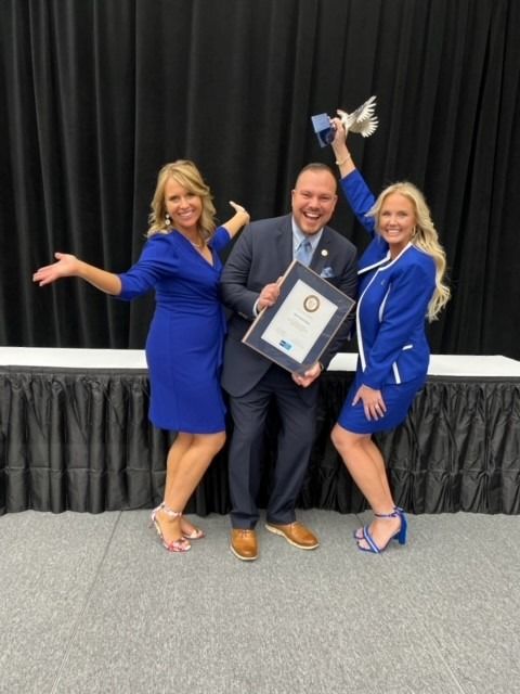 Three people celebrating an award. Man holds framed document, women flank him, one holds trophy. All are wearing blue.