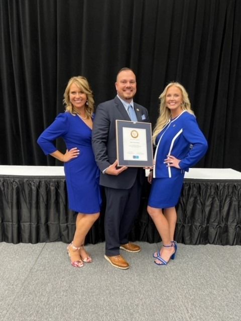 Three people in blue attire pose with an award, standing in front of a black backdrop.