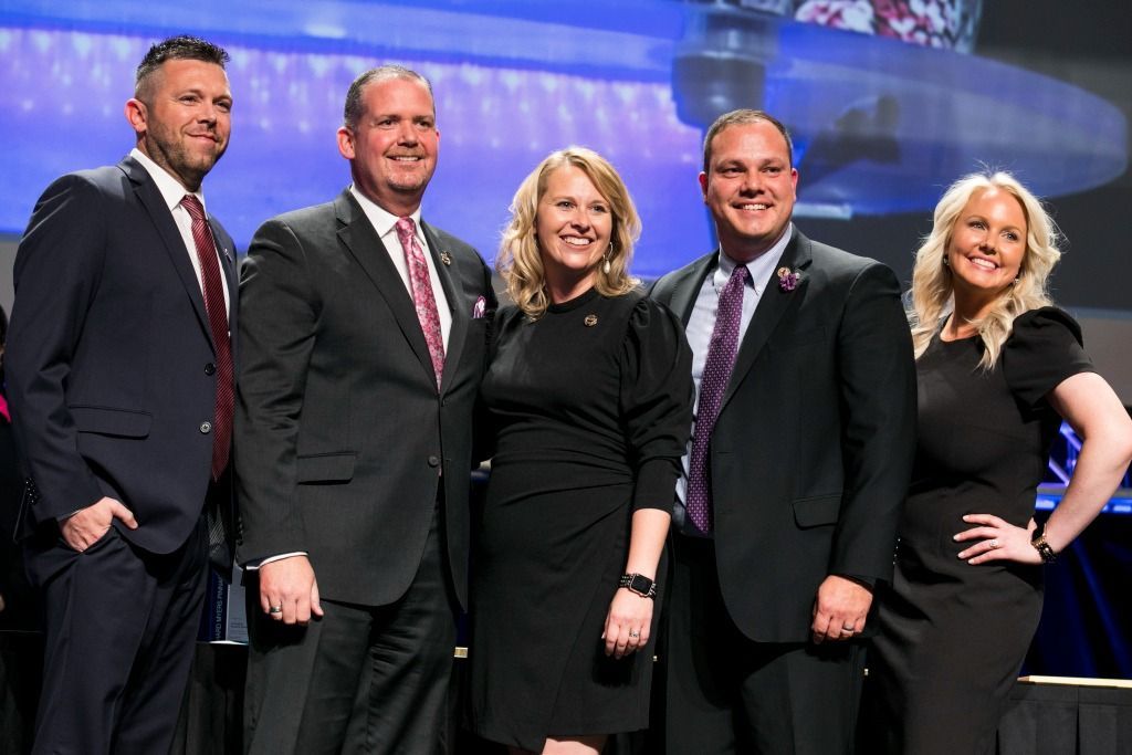 Five people in formal attire, posing on a stage with a screen in the background.