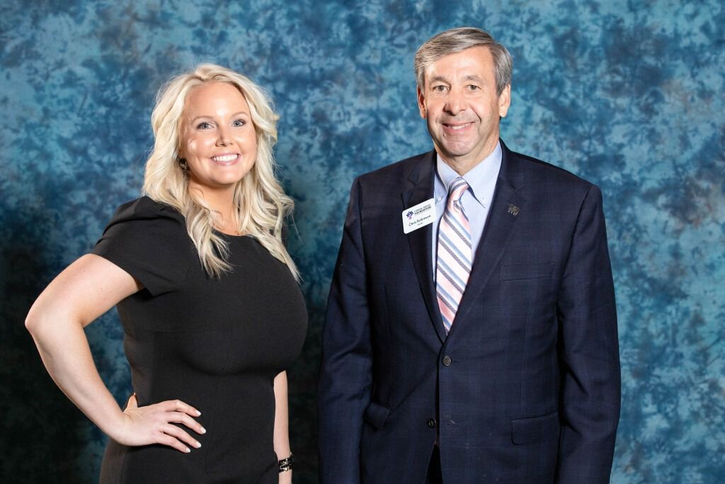 Woman in black dress and man in suit stand together in front of a blue backdrop.