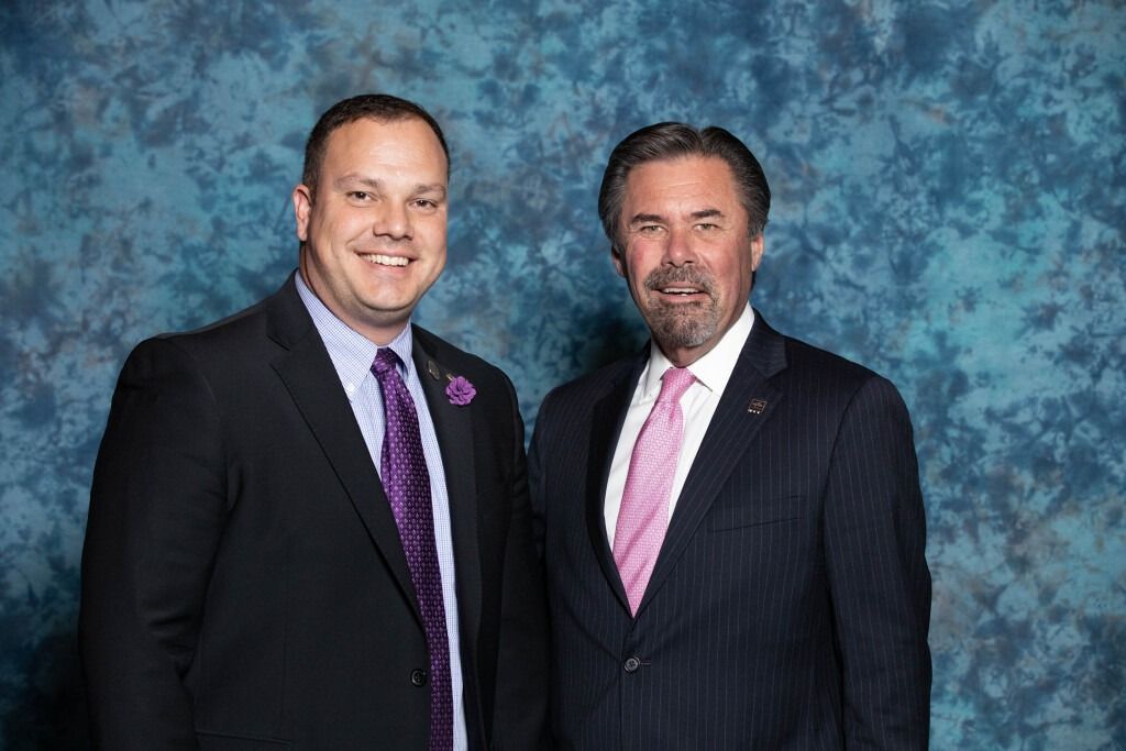 Two men in suits posing in front of a blue backdrop, one with a purple tie, the other with a pink tie.
