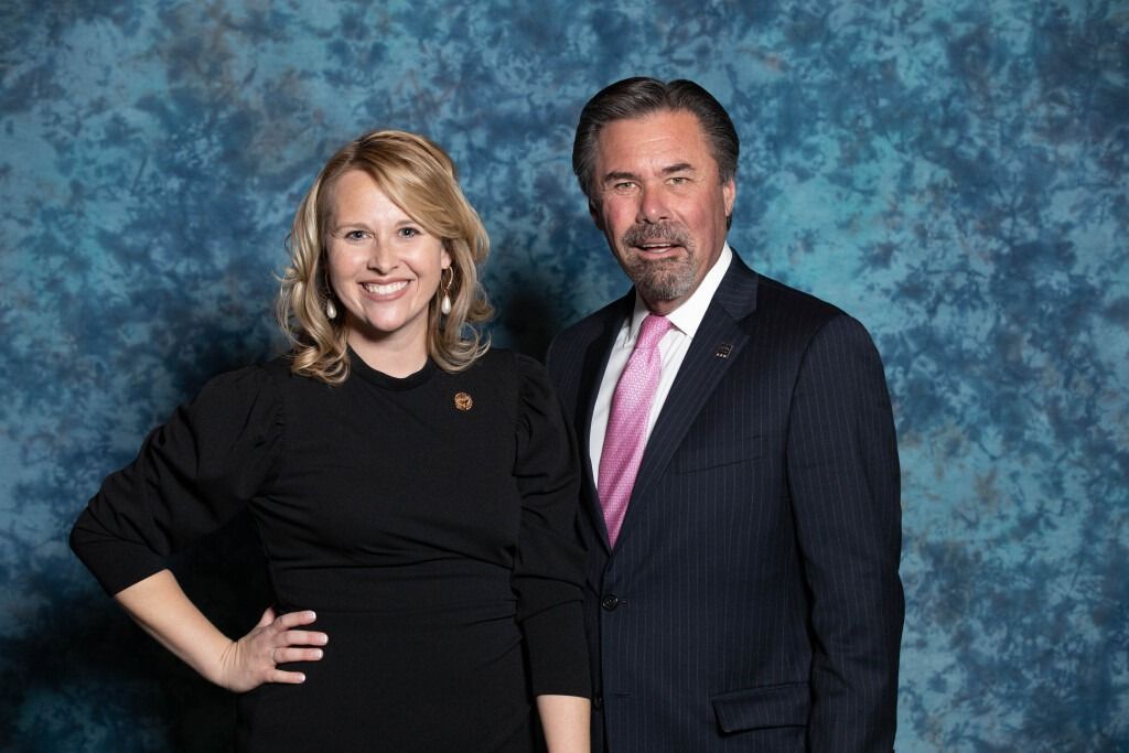 Woman in black dress with arm on hip and man in suit smiling in front of blue patterned backdrop.