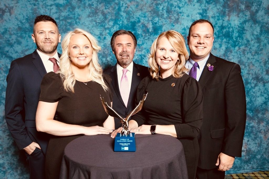 Group of five people around a table with a trophy. They are smiling, formal attire, against a blue backdrop.