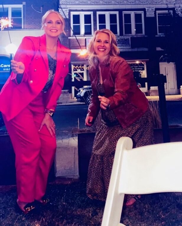 Two women smile, holding sparklers at an outdoor event, one in a pink suit, the other in a red jacket and floral dress.