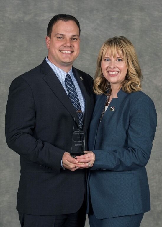 A man and woman in suits holding an award, smiling at the camera.