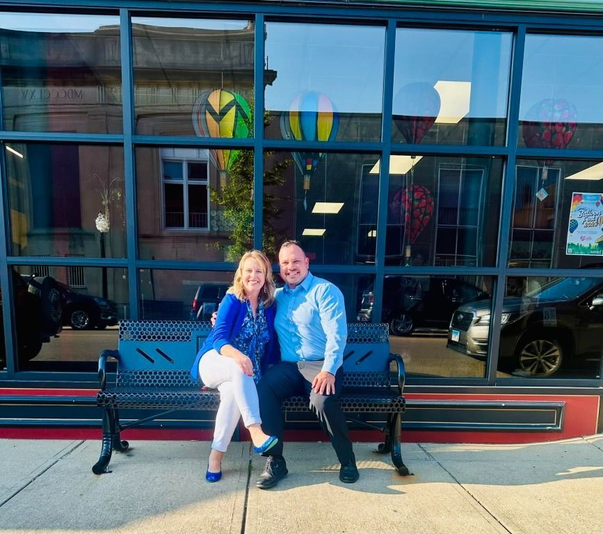 Couple seated on a bench, smiling, in front of a window with building reflections.