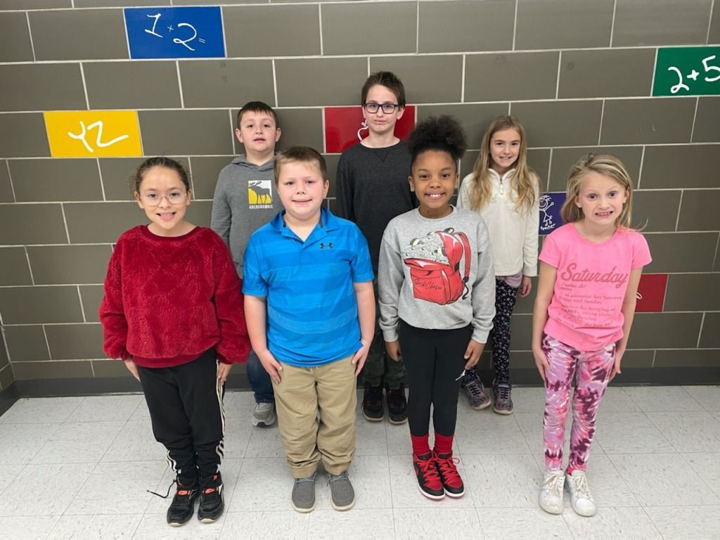 Seven children standing in front of a wall with colorful blocks with math problems.