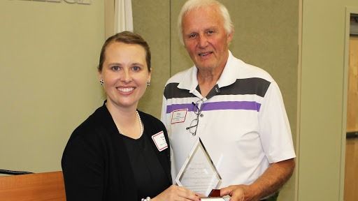 Woman presents an award to a man indoors, both smiling. The man wears a polo shirt, and the woman a black sweater.