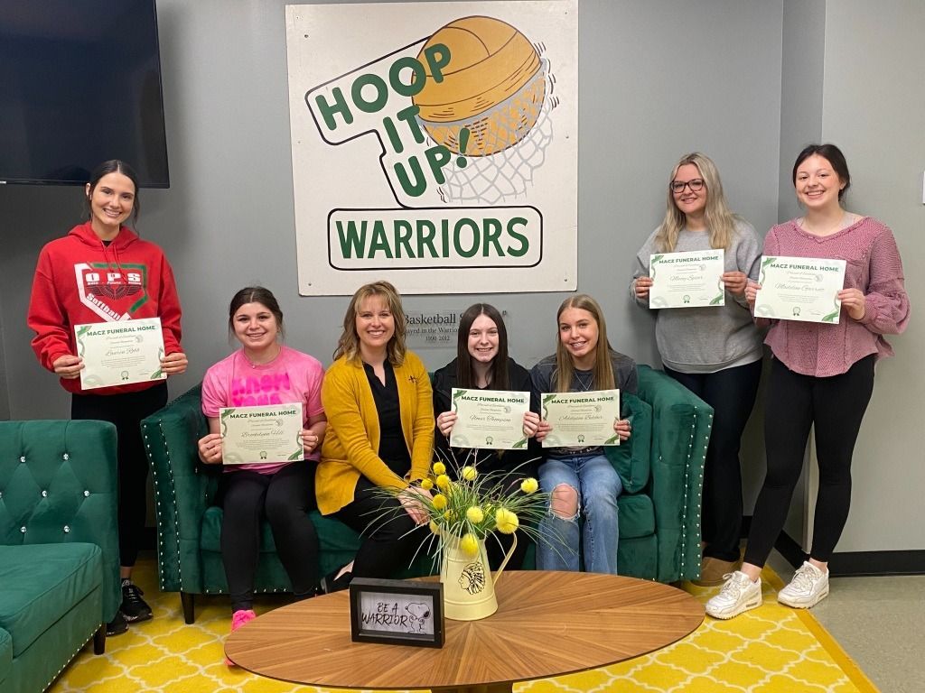 Group of people holding certificates, posing in front of a “Hoop It Up Warriors” banner. They are sitting on a green couch.