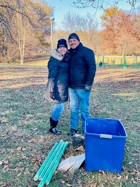 Couple smiling outdoors, with a blue bin, poles, and foliage.
