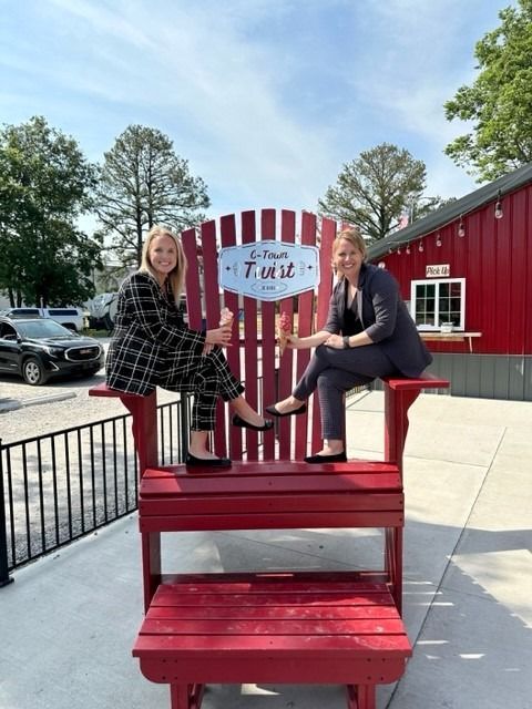 Two women sit on a giant red Adirondack chair. The chair has a sign that reads 